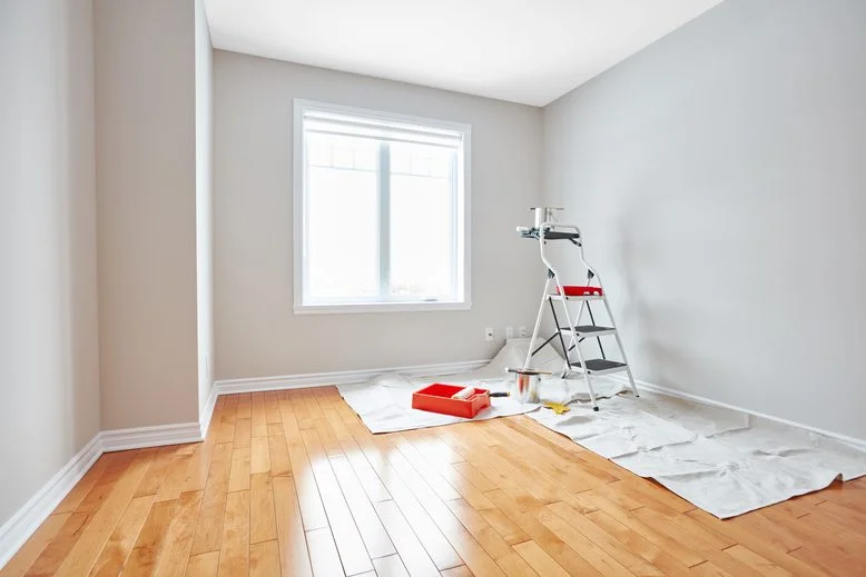A bright residential room undergoing interior painting, featuring a ladder, paint tray, and drop cloth on hardwood floors, managed by JD Local Builders in San Antonio, TX.